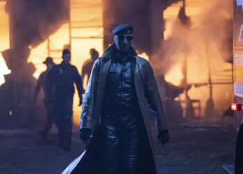Lee Pace as Evan McCone wearing a mask and trench coat standing in front of a burning building in The Running Man