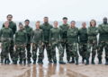 They stand on a beach, wearing green and camouflage military-style garb