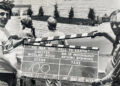 Two men, one on the left and another on the right, are holding a clapperboard, which is in the middle of this black and white photo.