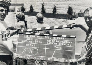 Two men, one on the left and another on the right, are holding a clapperboard, which is in the middle of this black and white photo.