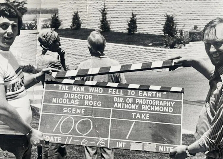 Two men, one on the left and another on the right, are holding a clapperboard, which is in the middle of this black and white photo.