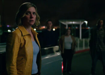 Rhea Seehorn's Carol Sturka standing beside a car with ominous figures on the background on Pluribus
