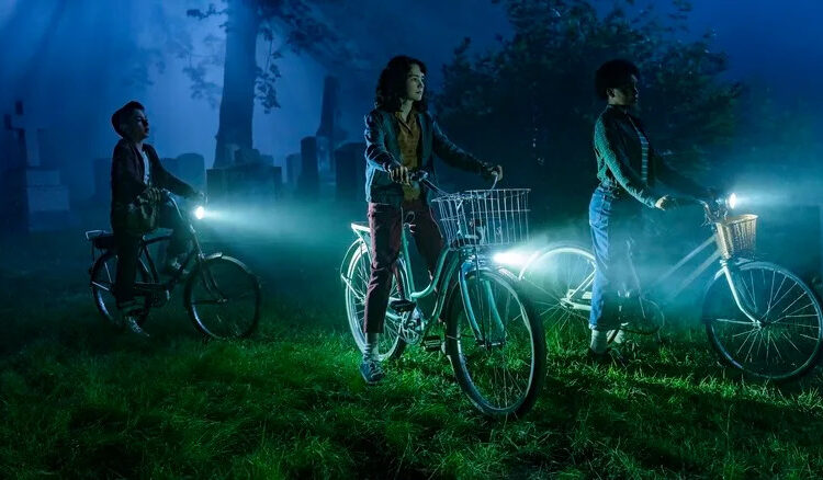 Arian S. Cartaya as Rich, Clara Stack as Lilly Bainbridge, and Amanda Christine as Ronnie Grogan riding bikes in a cemetery at night in It: Welcome to Derry