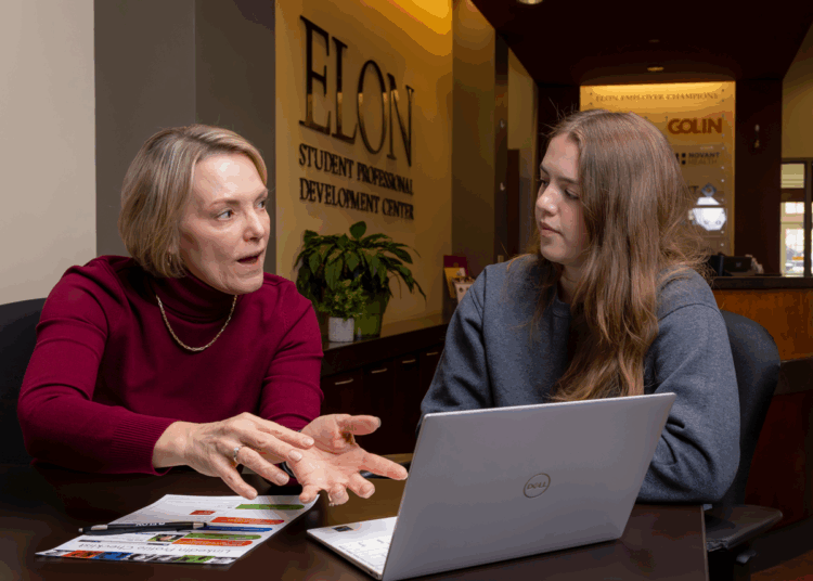 A career advisor speaks with a student inside the Elon Student Professional Development Center. The advisor gestures while explaining something, and the student listens attentively with an open laptop and documents on the table in front of them.