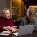 A career advisor speaks with a student inside the Elon Student Professional Development Center. The advisor gestures while explaining something, and the student listens attentively with an open laptop and documents on the table in front of them.