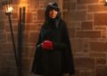 Claudia Winkleman wearing a black cloak and red gloves, smiling as she looks to the side.