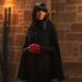 Claudia Winkleman wearing a black cloak and red gloves, smiling as she looks to the side.