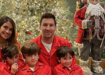A family of five wearing matching red pajamas sits in front of a decorated Christmas tree. Three children sit in front of two adults. A large Santa Claus decoration stands beside the tree.