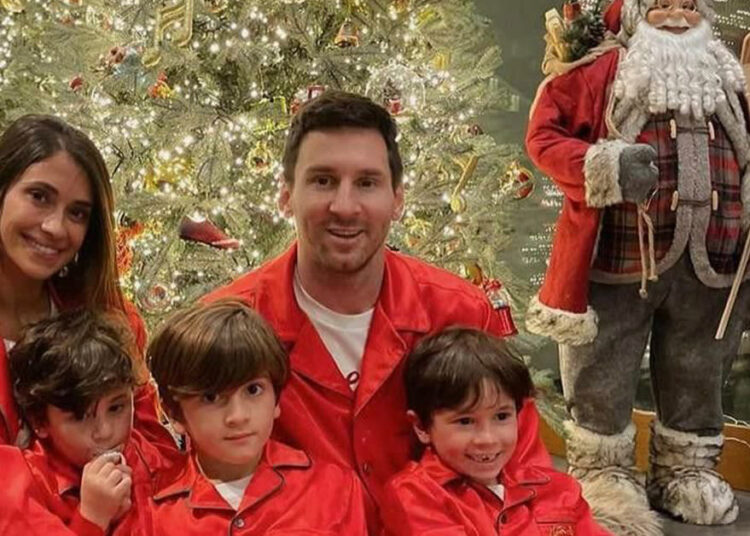 A family of five wearing matching red pajamas sits in front of a decorated Christmas tree. Three children sit in front of two adults. A large Santa Claus decoration stands beside the tree.