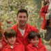 A family of five wearing matching red pajamas sits in front of a decorated Christmas tree. Three children sit in front of two adults. A large Santa Claus decoration stands beside the tree.