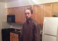 A man with light skin and brown hair pulled back stands in a kitchen with wooden cabinets, a white refrigerator, and a black stove. He is wearing a brown zip-up jacket and looking at the camera with a neutral expression.