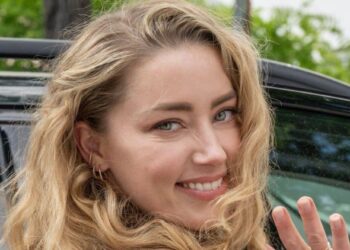 A woman with long, wavy blonde hair, wearing a tan dress and gold bracelet, smiles while turning toward the camera and waving, standing next to a black car outdoors.