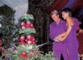 A smiling couple in matching purple outfits cut a large, colorful wedding cake with greenery and red apples on it. They are under a decorated tent, with lush drapery in the background.