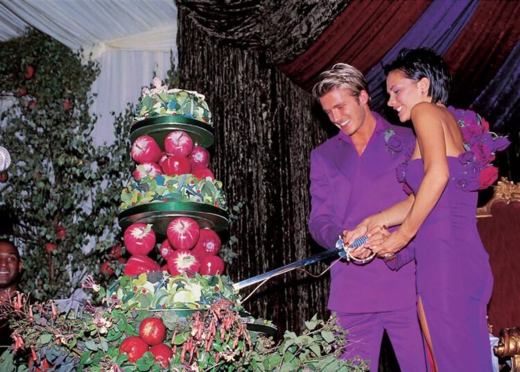 A smiling couple in matching purple outfits cut a large, colorful wedding cake with greenery and red apples on it. They are under a decorated tent, with lush drapery in the background.
