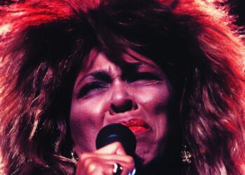 A person with big, wild hair and bright red lipstick sings passionately into a microphone on stage, eyes closed in emotion, with dramatic lighting highlighting their face.
