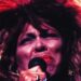A person with big, wild hair and bright red lipstick sings passionately into a microphone on stage, eyes closed in emotion, with dramatic lighting highlighting their face.
