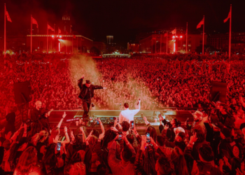 A massive crowd at a nighttime outdoor concert is bathed in red light as two DJs perform on stage with hands raised, many attendees filming with phones.