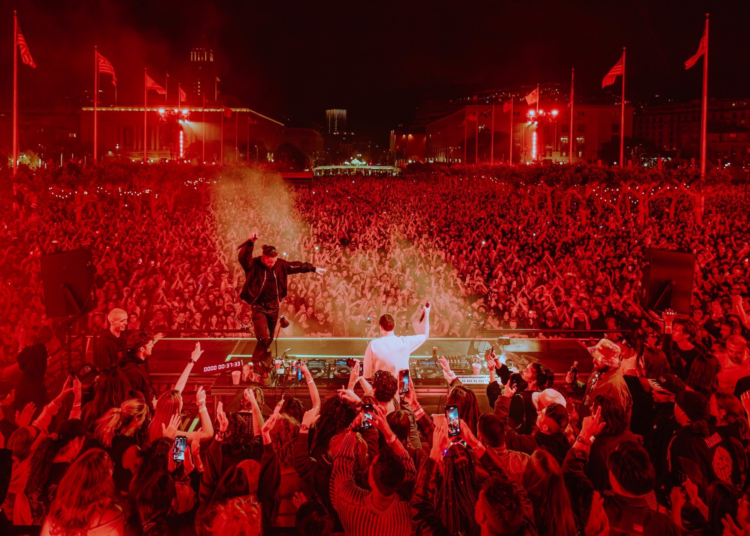 A massive crowd at a nighttime outdoor concert is bathed in red light as two DJs perform on stage with hands raised, many attendees filming with phones.