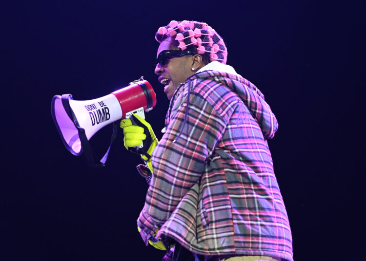 A person in a plaid jacket and pink curlers holds a megaphone labeled "DON'T BE DUMB" on stage, wearing sunglasses and gloves.