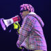 A person in a plaid jacket and pink curlers holds a megaphone labeled "DON'T BE DUMB" on stage, wearing sunglasses and gloves.