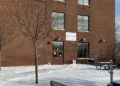 Brick building of three stories, attached to lower extension at left, is seen in snowy environment on a sunny day. Empty park benches appear in foreground.