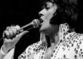 White man with shoulder-length dark hair sings into microphone. He wears a rhinestone-studded jumpsuit open at the neck, with medallion visible hanging from a chain around his neck.