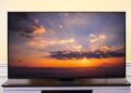 A Hisense U8QG Series flat-screen television sits on a dark media console in front of a light yellow wall. The screen displays a colorful sunset over a mountain range, with sunbeams radiating from behind clouds.