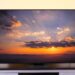 A Hisense U8QG Series flat-screen television sits on a dark media console in front of a light yellow wall. The screen displays a colorful sunset over a mountain range, with sunbeams radiating from behind clouds.
