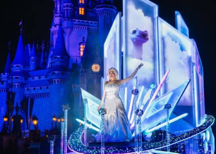 A performer dressed as Elsa from Frozen stands on an illuminated float with icy blue decorations, captivating Disney fans in front of a brightly lit castle at night during one of the magical Disney parades.