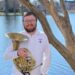A white man wears a white shirt and holds a French horn in an outdoor photo. Tyler Goodwin-Souffront.