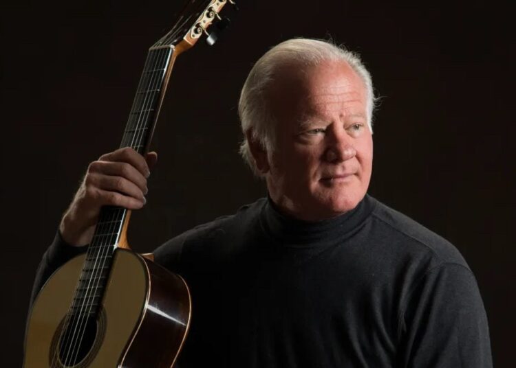 A white man with white hair wears a black shirt and holds an acoustic guitar. Gordon Kreplin