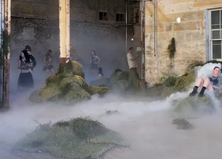 People gather around small grassed hills and fog in a sandstone industrial room