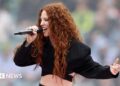 Jess Glynne is singing into a mic, she is wearing a cropped black suit jacket. SHe has long curly ginger hair.