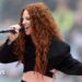 Jess Glynne is singing into a mic, she is wearing a cropped black suit jacket. SHe has long curly ginger hair.