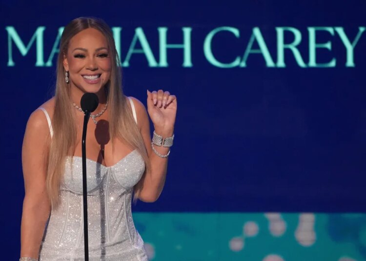 Mariah Carey smiling at a microphone with her name in the background.