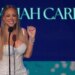 Mariah Carey smiling at a microphone with her name in the background.