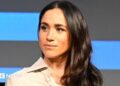 Meghan, who has long wavy brown hair, has a neutral expression as she looks on during a talk on stage
