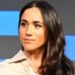 Meghan, who has long wavy brown hair, has a neutral expression as she looks on during a talk on stage