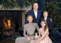 Princess Charlene wears a long taupe dress with a pleated skirt, long sleeves, and a turtle neck, and poses next to a burning fireplace and a Christmas tree with her pet dog, her twin children, Princess Gabriella and Prince Jacques, and husband Prince Albert