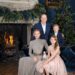 Princess Charlene wears a long taupe dress with a pleated skirt, long sleeves, and a turtle neck, and poses next to a burning fireplace and a Christmas tree with her pet dog, her twin children, Princess Gabriella and Prince Jacques, and husband Prince Albert