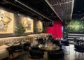 The dining room of a restaurant with beige booths along the wall under carved wooden artwork. Tables and chairs are placed around the room. A large greenery-covered sign says 'Tamasha' behind a DJ booth.