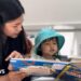 A mother and her daughter read a book. The mother has long black hair, and is wearing a brown/rust coloured top. She is holding a book that her young daughter appears to be reading. The child has shoulder length dark hair, and is wearing a white top and a turquoise sun hat. There is a book in the foreground, with the word "dinosaur" visible. A blue sofa is to the centre right of the image, and book shelves to the left.