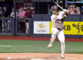 A baseball player in a white uniform with a maroon helmet and a bat in hand, standing at home plate, with "KB" and "KB LIFE" visible on the surrounding signage.
