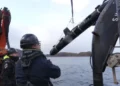 BBC Three men in hardhats onboard a ship launching  a SG-1 Fathom glider into the sea. It is lifted by a pulley and lowered into the water