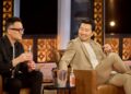 Two men sit next to each other in club chairs, laughing.