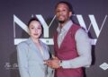 Two people posing in front of a black backdrop with large “NYFW” text and smaller sponsor logos