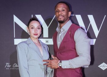 Two people posing in front of a black backdrop with large “NYFW” text and smaller sponsor logos