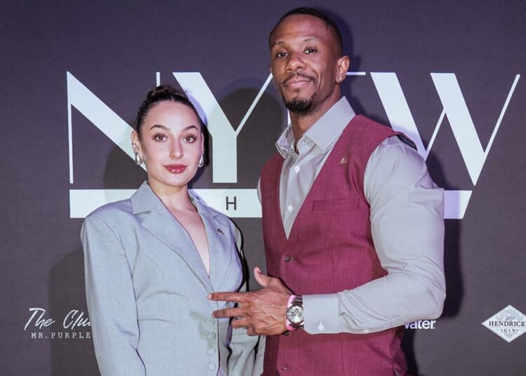Two people posing in front of a black backdrop with large “NYFW” text and smaller sponsor logos