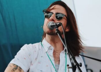 A close-up outdoor photo of a long-haired individual singing into a microphone. The subject is wearing dark sunglasses and a short-sleeved white button-down shirt with a small pattern. A tattoo is visible on their arm.