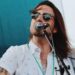A close-up outdoor photo of a long-haired individual singing into a microphone. The subject is wearing dark sunglasses and a short-sleeved white button-down shirt with a small pattern. A tattoo is visible on their arm.